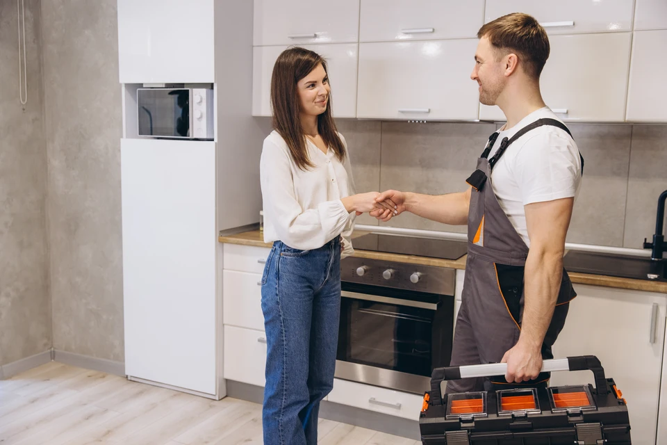 Photographie d'un professionnel de la rénovation intérieure, en tenue de travail (gilet ou chemise pratique), discutant avec un client devant des plans d'aménagement de cuisine ou de salle de vie étalés sur une table, dans un environnement intérieur résidentiel en cours de rénovation ou aménagement. L'image doit transmettre une relation de confiance, d'expertise et de collaboration entre l'artisan et son client, dans un contexte authentique d'accompagnement de projet.