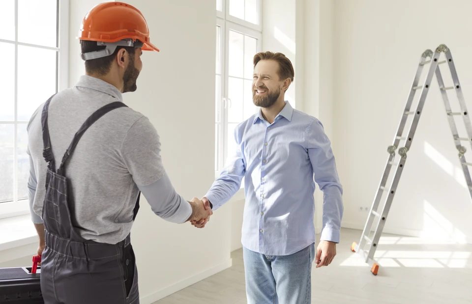 Photographie d'un artisan menuisier-agenceur dans son atelier ou sur un chantier de r&eacute;novation int&eacute;rieure, en interaction directe avec un client (conversation, explication, montage des plans ou pr&eacute;sentation de mat&eacute;riaux). L'artisan pr&eacute;sente des solutions d'agencement ou discute du projet avec le client dans un contexte professionnel authentique, illustrant le suivi personnalis&eacute; et le contact direct tout au long du projet de r&eacute;novation.